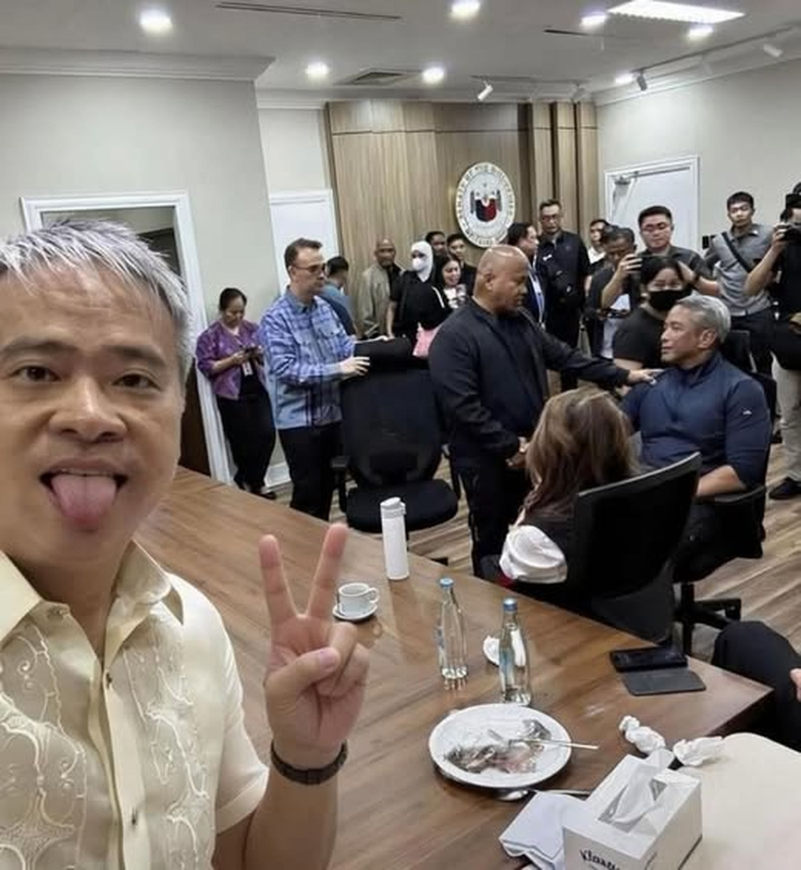 Un senador filipino saca la lengua para mofarse del pueblo mientras se celebra el triunfo del simulacro reciente en el senado filipino junto con un miembro del gabinete.  Destaca la calvicie del fugitivo Rolando Dela Rosa en el centro mientras habla con el miembro del gabinete del actual presidente de la Republica