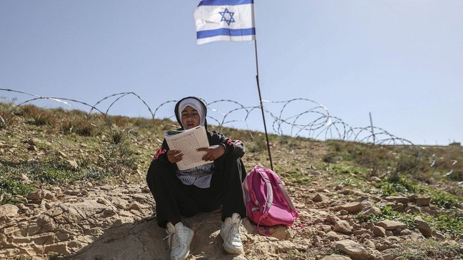 Niña palestina lee su libro de texto frente a una valla de alambre de púas.